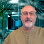 A middle-aged man with glasses and a trimmed gray beard is standing in front of a geometric, futuristic background.